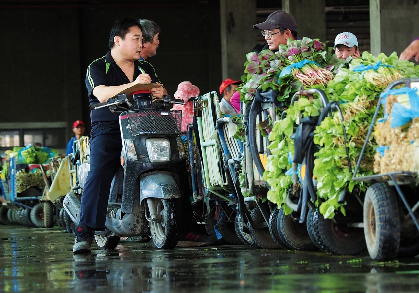 A buyer rides a small scooter among vegetable sellers as he places orders.